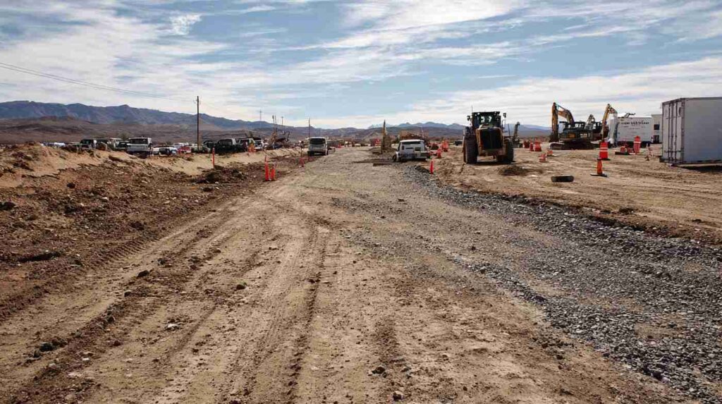 A dusty desert construction site with gravel being compacted for a new roadbed, supplied by an aggregate supplier near Barstow.