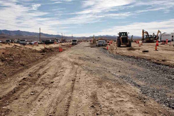 A dusty desert construction site with gravel being compacted for a new roadbed, supplied by an aggregate supplier near Barstow.